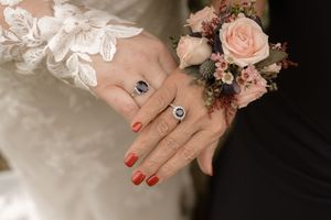 Close-up of two hands wearing matching sapphire rings with a floral wrist corsage on wedding day.