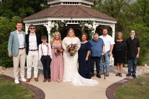 Wedding party poses in front of white gazebo adorned with flowers, bride in lace gown holds bouquet surrounded by family.