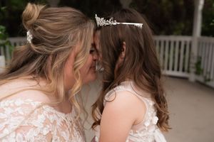 Bride and flower girl sharing a sweet forehead kiss, both wearing white lace dresses and tiaras at a wedding.