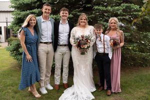 Wedding party poses outdoors with bride in lace gown holding bouquet surrounded by family members in formal attire.