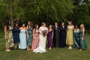 Bride in white gown stands smiling with large bridal party in colorful mismatched dresses outdoors.