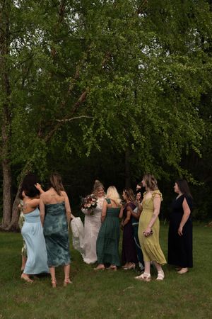 Bridesmaids in colorful mismatched dresses surround a bride holding a bouquet outdoors near lush green trees.