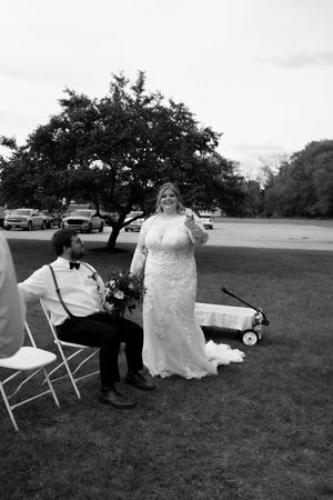 Black and white photo of bride in lace gown standing beside seated groom on lawn at wedding.
