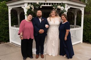 Wedding party poses before white gazebo with floral arch, bride in lace gown flanked by groom and two women in navy blue.