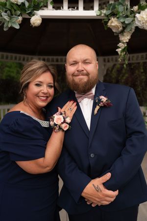 A woman and bald man in navy blue wedding attire pose together near a floral gazebo.