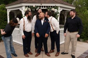 Groom in navy suit poses with groomsmen in white shirts and suspenders near a white gazebo at an outdoor wedding.