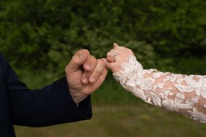 A bride in lace wedding dress and groom in navy suit gently hold pinkies outdoors with green foliage background.