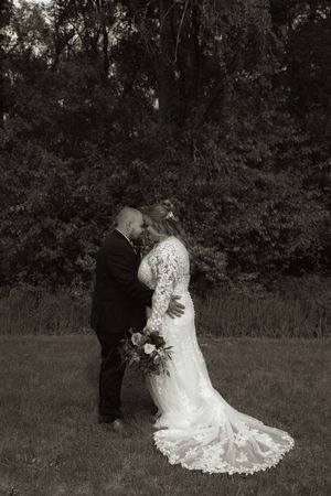Black and white wedding photo of bride in lace gown and groom in suit sharing intimate moment outdoors.