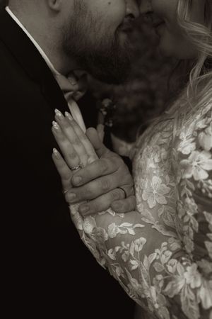 Black and white close-up of bride and groom kissing with wedding rings visible on clasped hands.