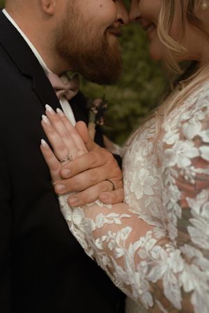 Close-up of bride and groom's hands showing wedding rings, bride wearing a floral lace long-sleeve gown.