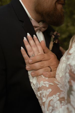 Close-up of bride and groom hands showing wedding rings, bride wearing lace gown and groom in black suit.