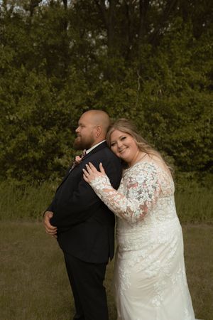 Bride in lace gown embraces groom in black suit from behind in a lush green outdoor setting.