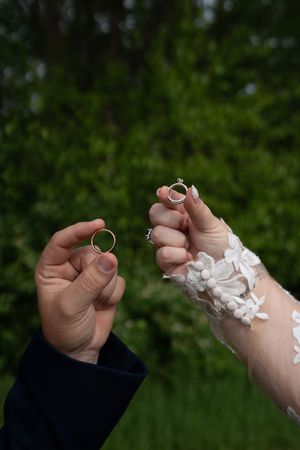 Bride and groom holding wedding rings up together outdoors with green trees in background.