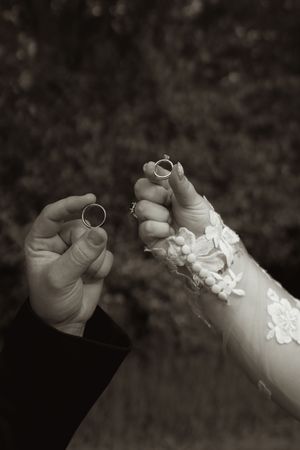 Black and white photo of bride and groom holding wedding rings up toward camera, bride wearing lace wedding dress.