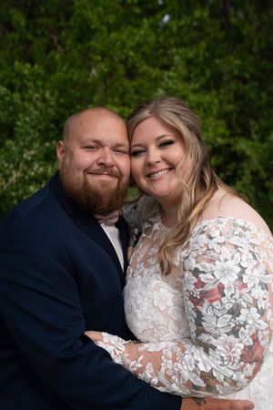 Smiling bride and bearded groom embrace closely for a portrait surrounded by green foliage.
