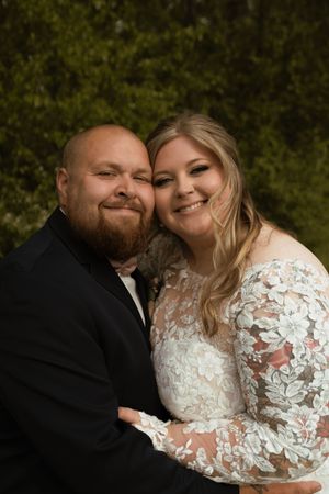 Smiling bride and groom in lace dress and black suit pose together at outdoor wedding.
