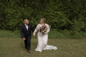 Bride in lace gown and groom in navy suit walk hand in hand across a green grassy field.