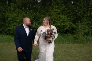 Smiling bride and groom walk hand in hand through a green meadow, bride holding a colorful bouquet.