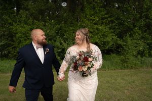 Bride in lace gown and groom in navy suit hold hands walking through green field.