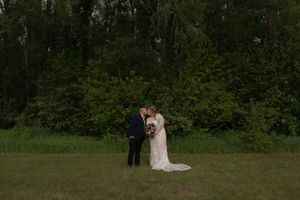 Bride and groom share a kiss in a green meadow surrounded by tall trees during outdoor wedding portraits.