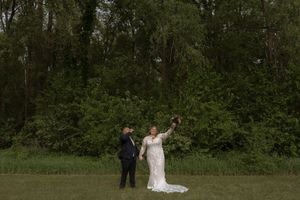 Bride in lace gown raises bouquet while groom watches during outdoor wedding portraits in a grassy field.