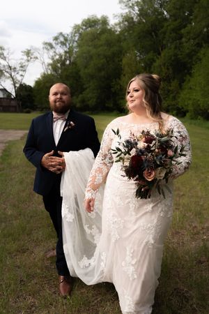 A groom in navy suit carries the train of his bride's lace gown as they walk outdoors.