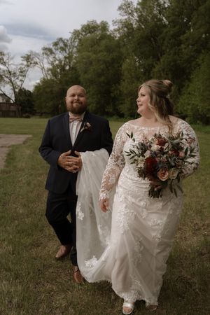 Bride in lace gown holding bouquet walks with groom in navy suit on grassy path surrounded by trees.