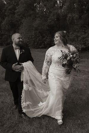 Black and white photo of bride and groom walking outdoors, bride holding bouquet in lace gown, groom in suit carrying train.