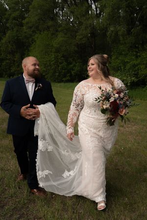 Bride in lace gown holding floral bouquet walks with groom in navy suit through green meadow.