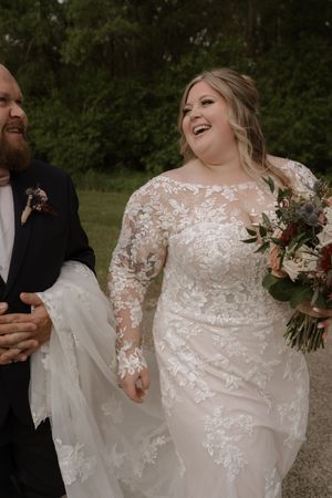 Laughing bride in lace long-sleeve wedding gown walks with bearded groom in black tuxedo holding bouquet.