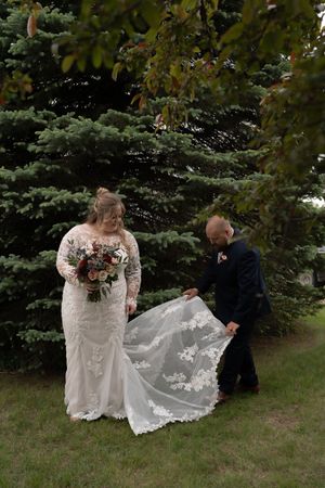 Groom in navy suit adjusts bride's lace wedding gown train near evergreen tree outdoors.
