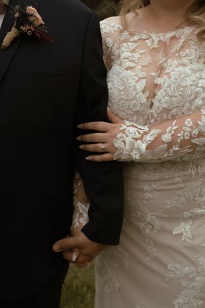 Close-up of bride in lace gown holding grooms arm showing diamond engagement ring and boutonniere.