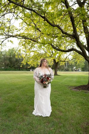 Bride in long-sleeve lace wedding dress holds floral bouquet beneath a leafy tree on green lawn.