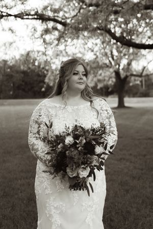 Black and white portrait of a bride in lace gown holding a bouquet beneath a large tree.