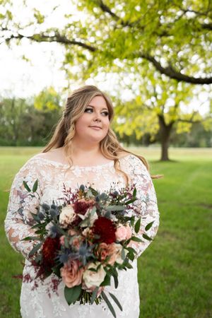 Bride in lace long-sleeve gown holding colorful bouquet of roses outdoors under green tree.