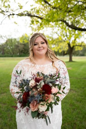 Bride in lace long-sleeve wedding gown holding bouquet of burgundy, blush, and dusty blue flowers outdoors.