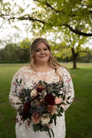 Smiling bride in long-sleeve lace wedding gown holding lush bouquet of burgundy and blush flowers under tree.
