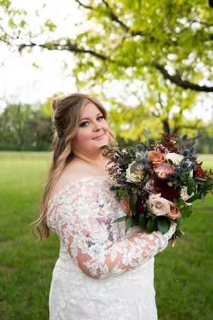 Bride in lace long-sleeve gown smiling over shoulder while holding a lush bouquet of burgundy and blush blooms.