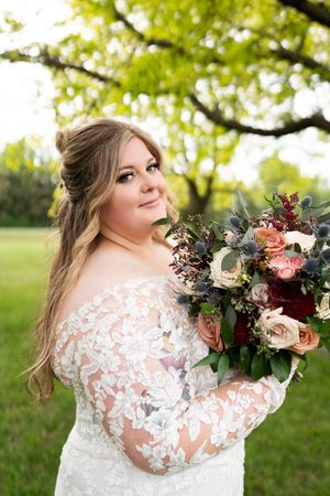 Bride in lace off-shoulder gown smiling over shoulder holding bouquet of roses and greenery.