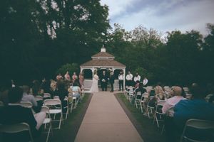 Outdoor wedding ceremony at dusk beneath a white gazebo with guests seated in white folding chairs.