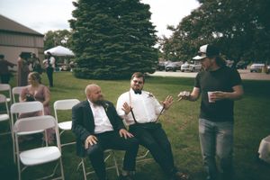 Groom in tuxedo and groomsman in suspenders laugh and chat with a guest at outdoor reception.