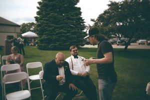 Groomsmen in tuxedo and suspenders converse with a guest in a cap at an outdoor wedding.