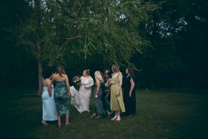 Bride in white gown stands with bridesmaids in colorful dresses under a weeping tree at outdoor wedding.