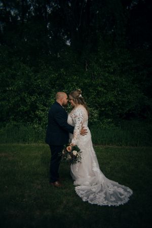 Bride in floral lace gown with bouquet and groom embrace at dusk in lush garden setting.