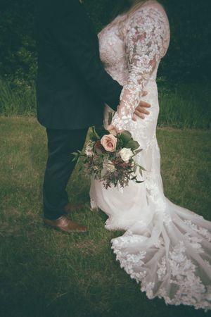 Bride in lace long-sleeve wedding gown holding floral bouquet stands with groom in dark suit on green lawn.