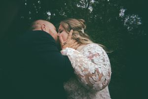 Bride in lace gown and groom share a romantic kiss surrounded by dark lush trees outdoors.