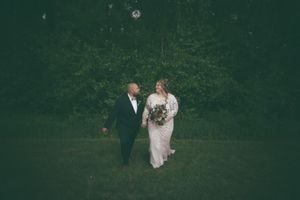 Bride holding bouquet and groom in navy suit walk together through a lush green outdoor setting.