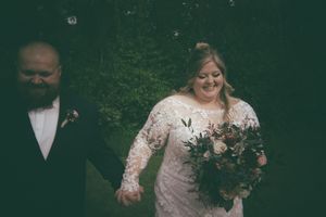 A smiling bride in lace gown holds a bouquet while walking hand in hand with her groom.