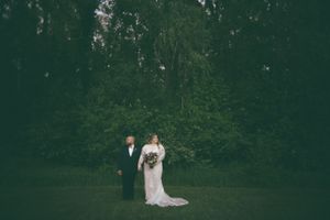 Bride in white gown holds bouquet beside escort in lush green outdoor garden setting at wedding ceremony.