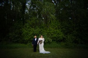 Bride in long lace gown and escort stand together in front of tall green trees at dusk.